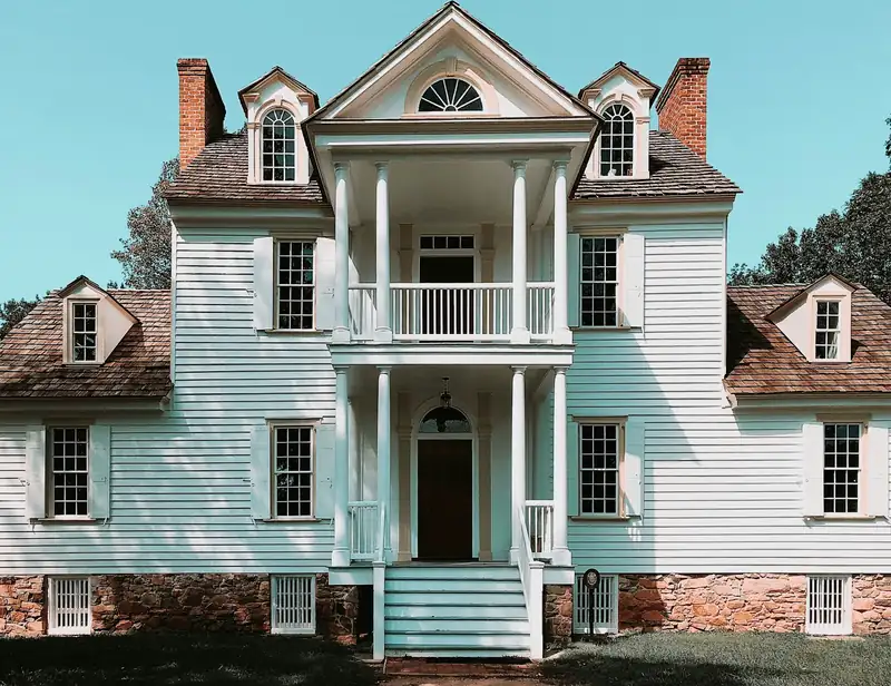 Casa de dos pisos pintada de blanco, con balcón y columnas, bajo un cielo azul